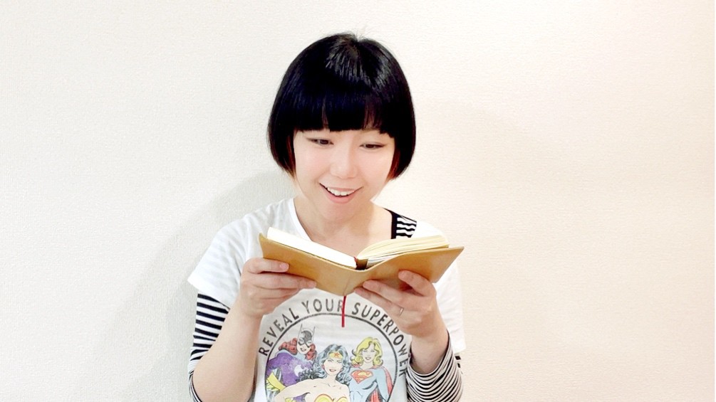 読書をしているおかっぱミユキ。革製のブックカバーがついている文庫本を書か手で持ち、面白そうな表情をしている。
