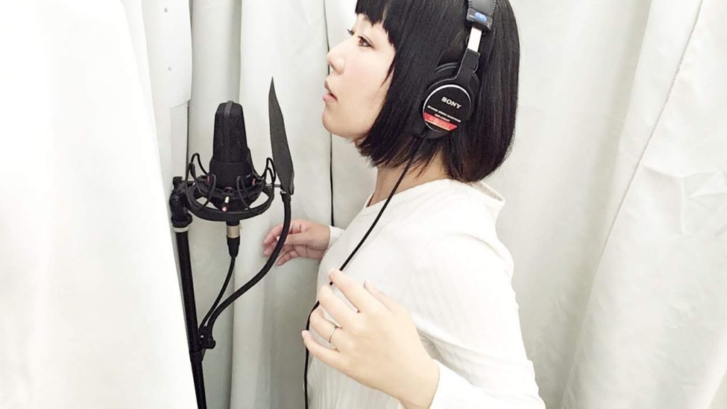 歌のレコーディングをするおかっぱミユキの写真。ヘッドフォンをつけて、コンデンサーマイクの前に立っている。