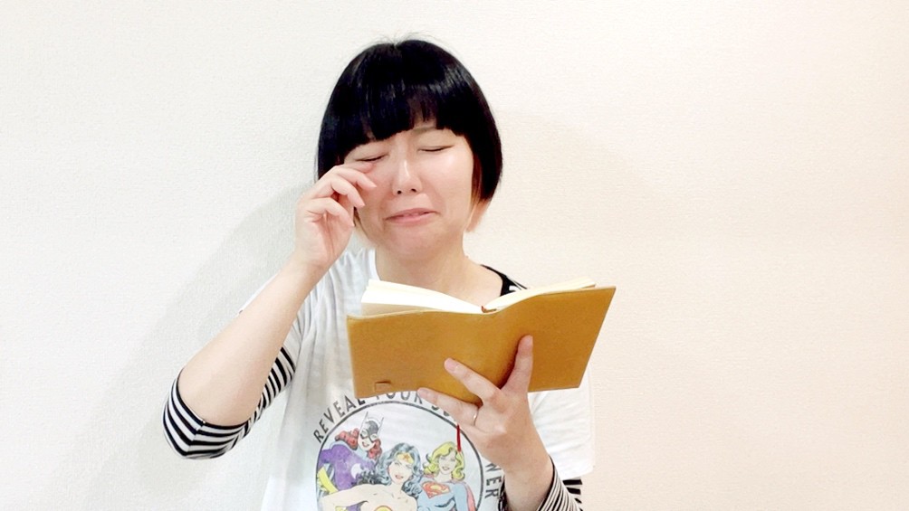 読書をしているおかっぱミユキ。革製のブックカバーがついている文庫本を書か手で持ち、悲しそうな表情をしている。