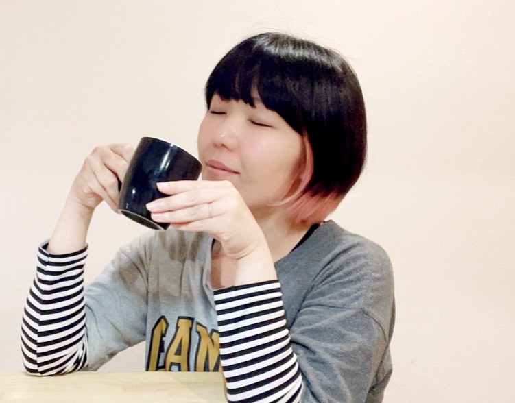 コーヒーを楽しむおかっぱミユキの写真。黒いマグカップを手に、ニコニコしている。