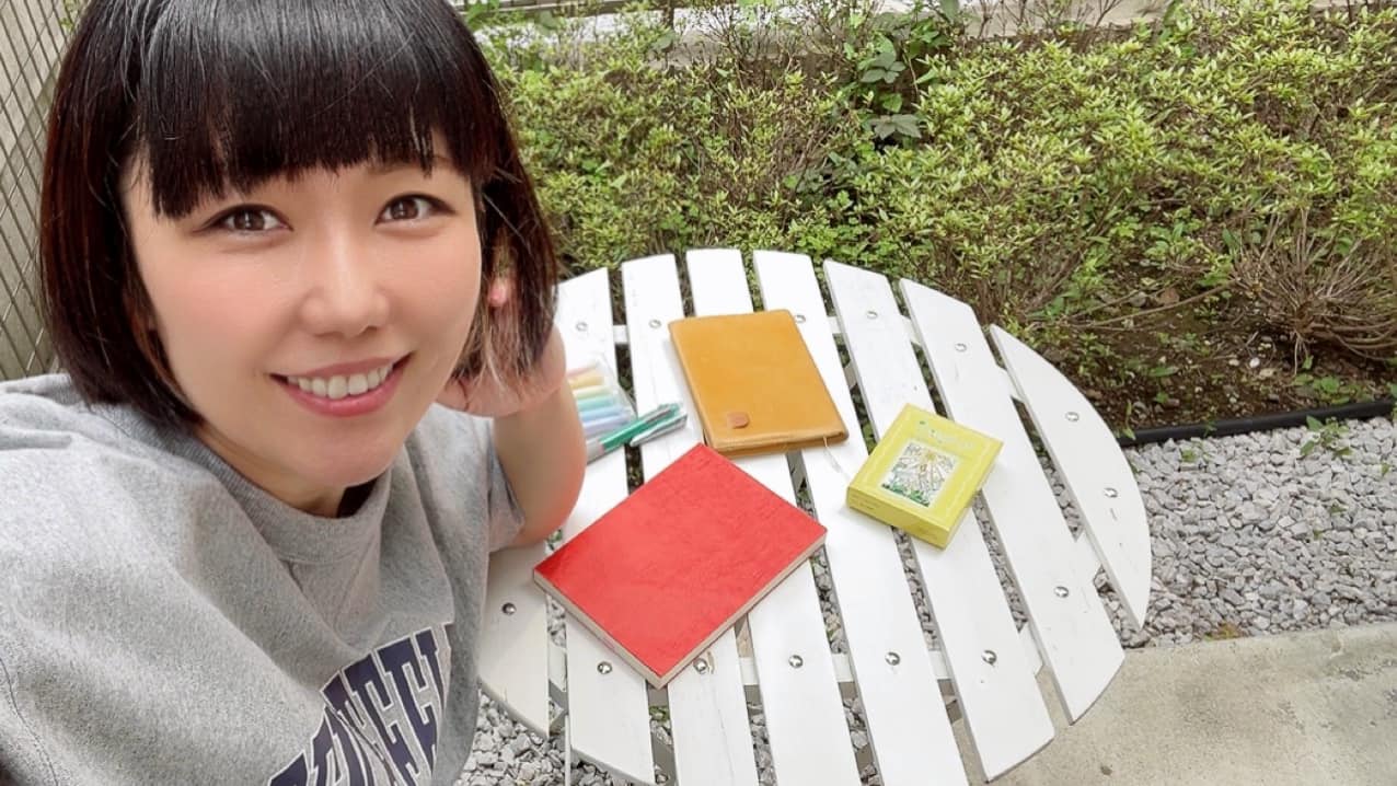 庭で微笑むおかっぱミユキの写真。ガーデンテーブルの上に手帳や読みかけの本が置いてある。