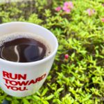 マグカップに入ったブラックコーヒーの写真。背景には咲き掛けのサツキが写っている。