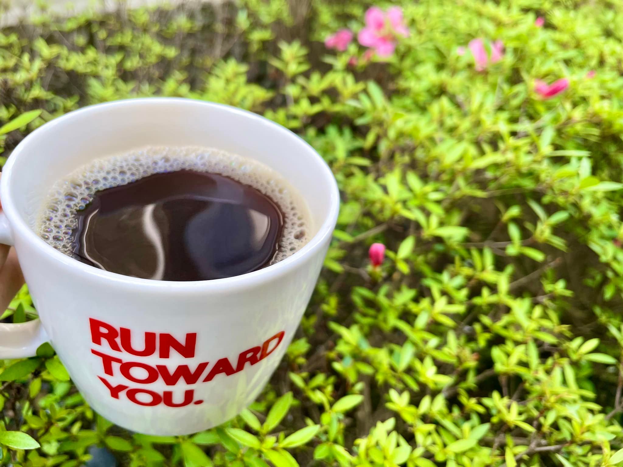 マグカップに入ったブラックコーヒーの写真。背景には咲き掛けのサツキが写っている。