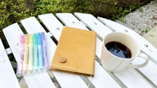 ガーデンテーブルにコーヒーと手帳、色とりどりのペンが並んでいる写真。奥には緑が茂っていて、柔らかい光が差している。