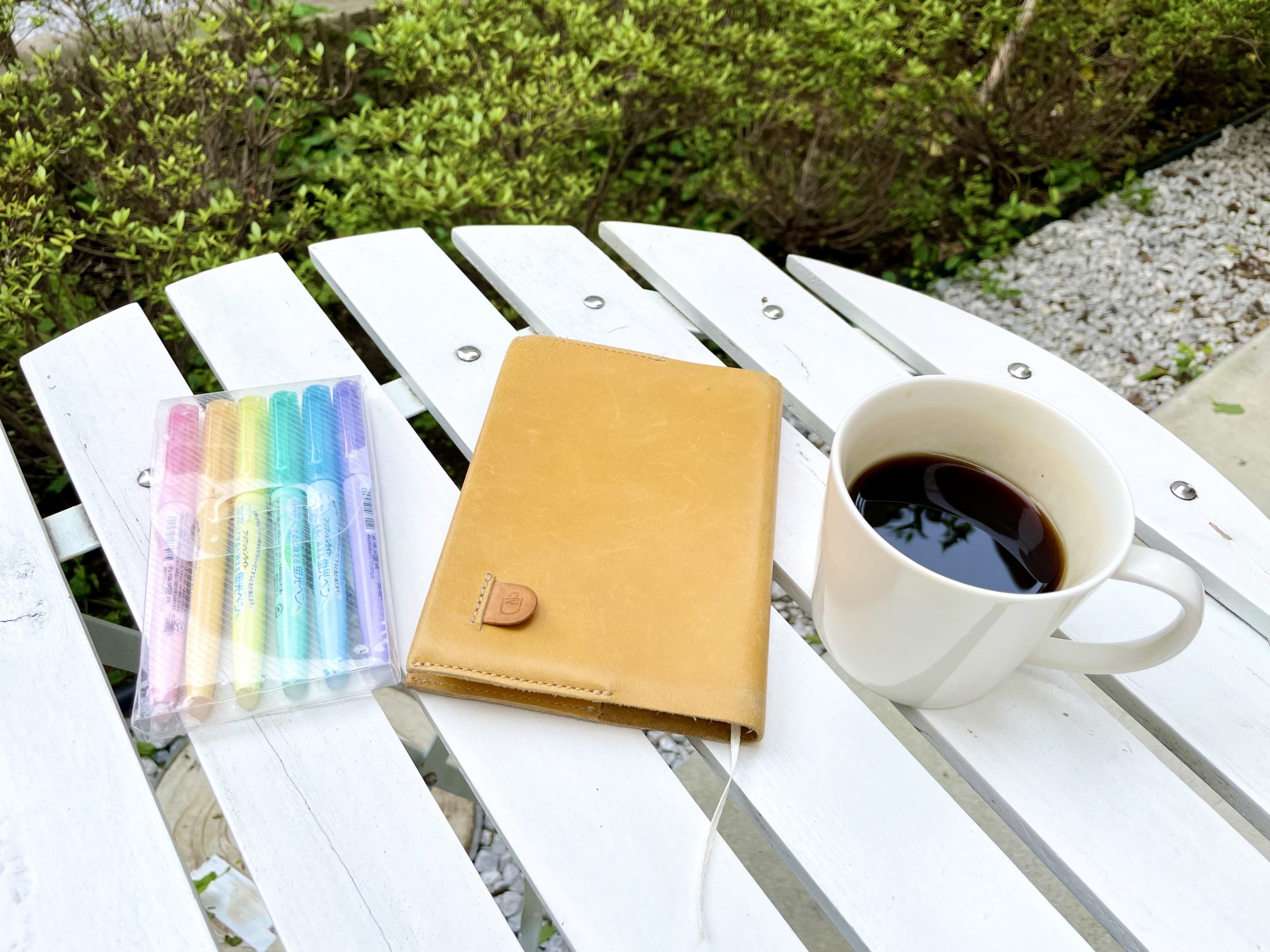 ガーデンテーブルにコーヒーと手帳、色とりどりのペンが並んでいる写真。奥には緑が茂っていて、柔らかい光が差している。