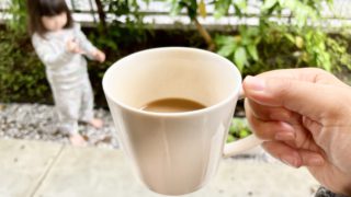 手に持たれているコーヒーカップの写真。奥には、子供が遊ぶ姿がぼんやりと写っている。