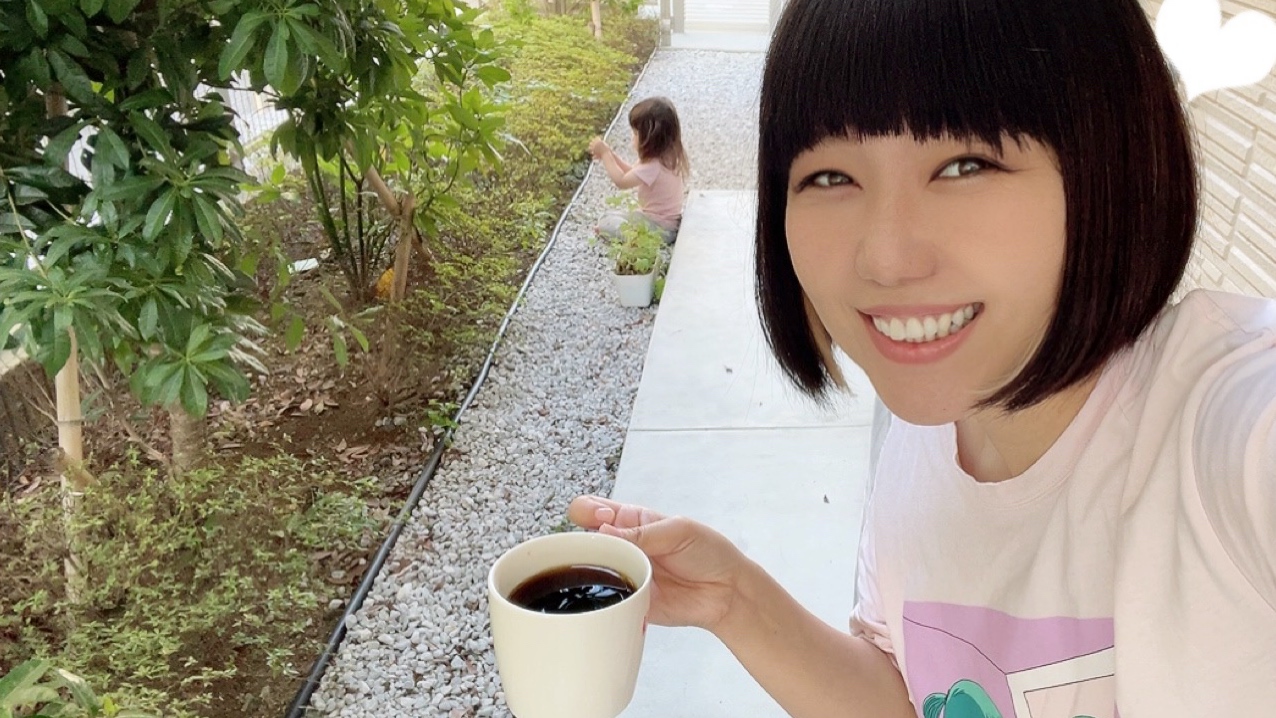 コーヒーカップを片手に微笑むおかっぱミユキの写真。自宅の庭で子供が遊んでいる。