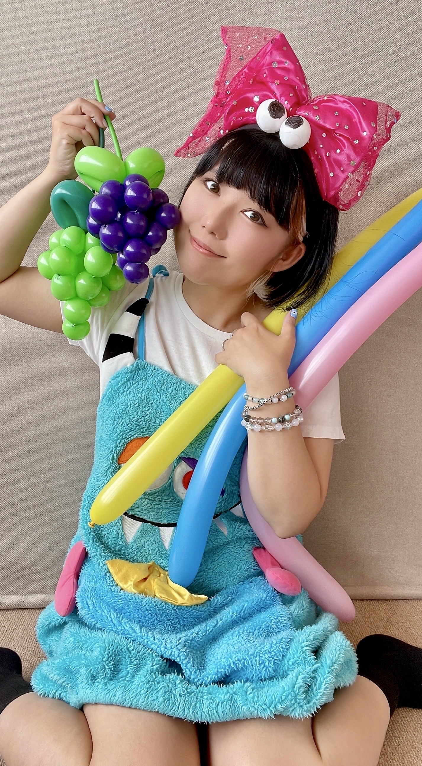 おかっぱミユキの写真。葡萄（ぶどう）などのカラフルなバルーンアートを両手に持ち、ニコッと笑っている。
