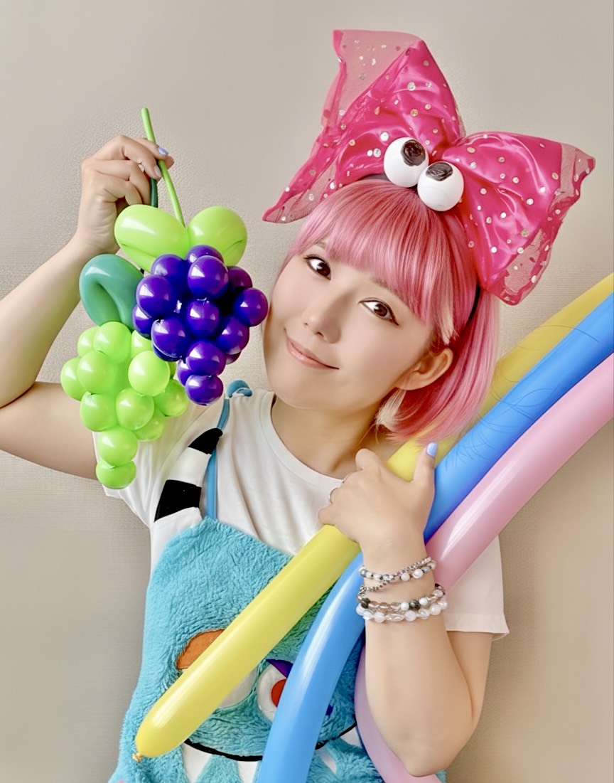 ピンク色の髪のおかっぱミユキが、カラフルなバルーンアートを抱えてにっこり微笑んでいる写真